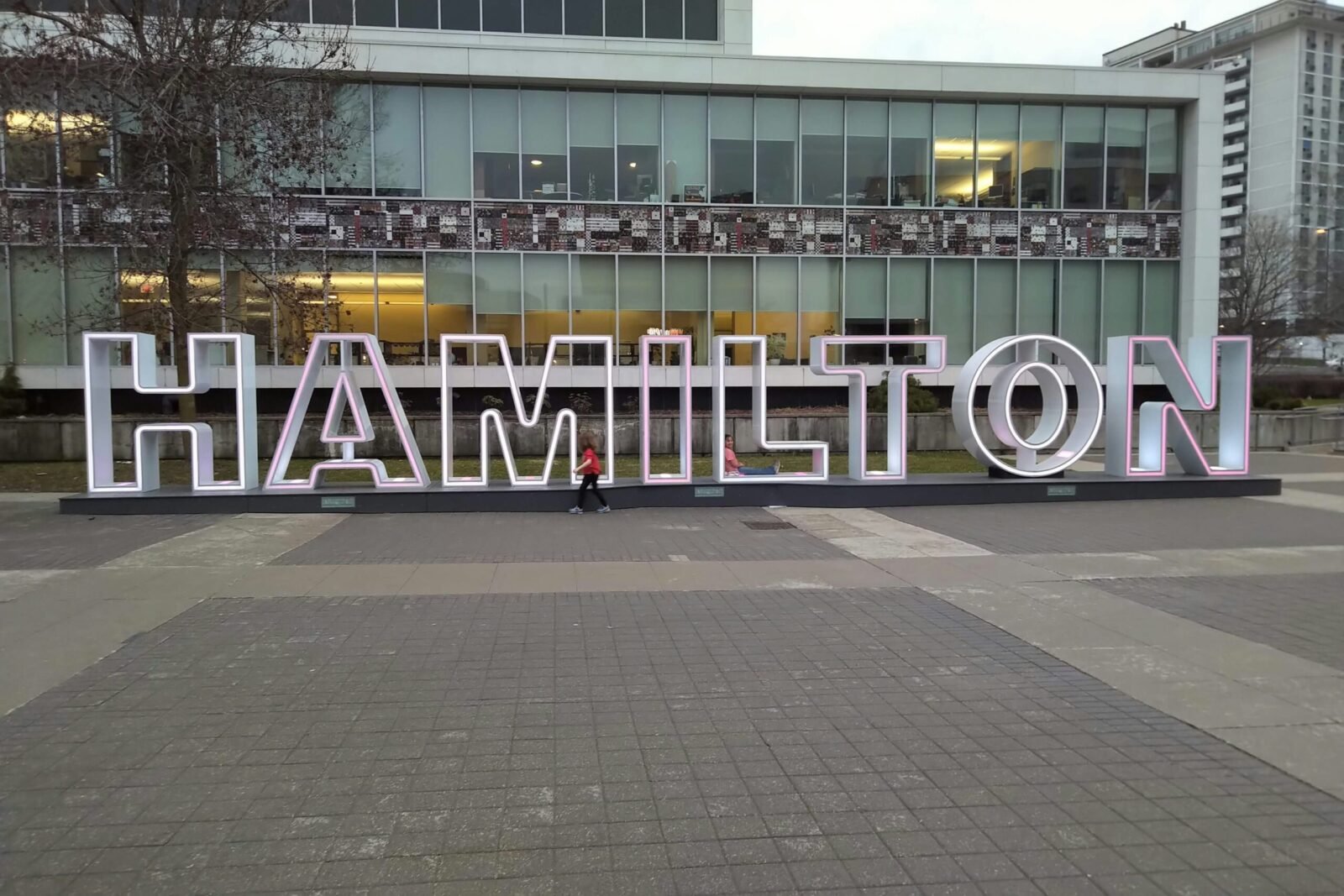 A large illuminated 3D Hamilton city sign in a public downtown plaza in front of a modern glass building