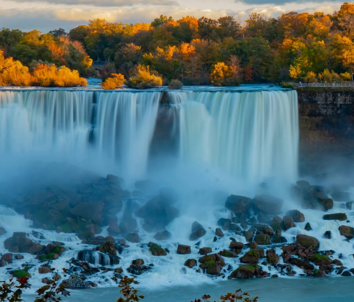Long exposure of the American Falls framed by golden autumn trees, with water flowing over giant boulders.